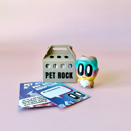 A colorful figure with big eyes and a heart stands beside a gray carrier labeled "PET ROCK" and adoption cards.