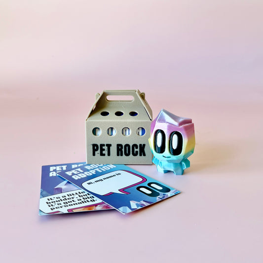 A colorful, cartoonish rock character stands beside a gray carrier and adoption cards on a soft pink background.