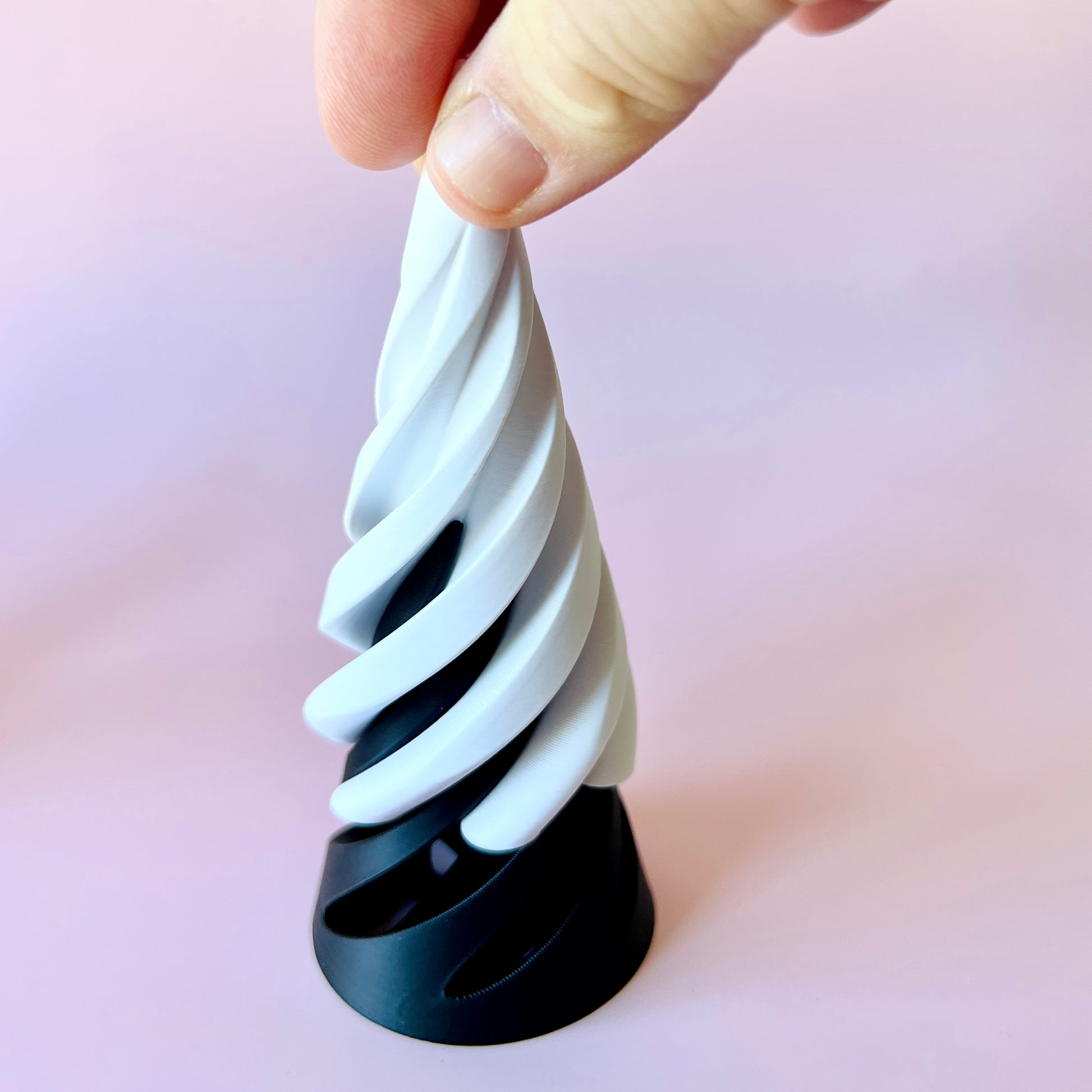 A person holds a spiraled, dual-colored structure with a black base against a soft pink background.