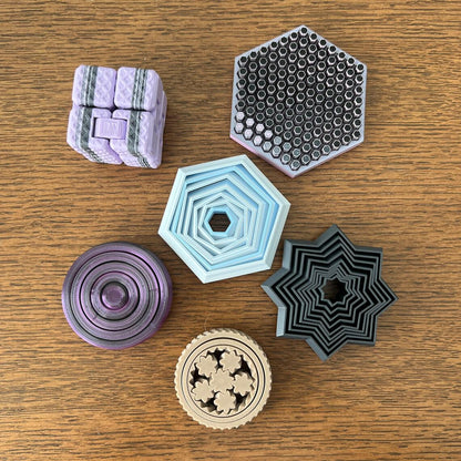 Colorful 3D-printed fidget shapes: a cube, hexagons, a star, and a round design, all on a wooden surface.