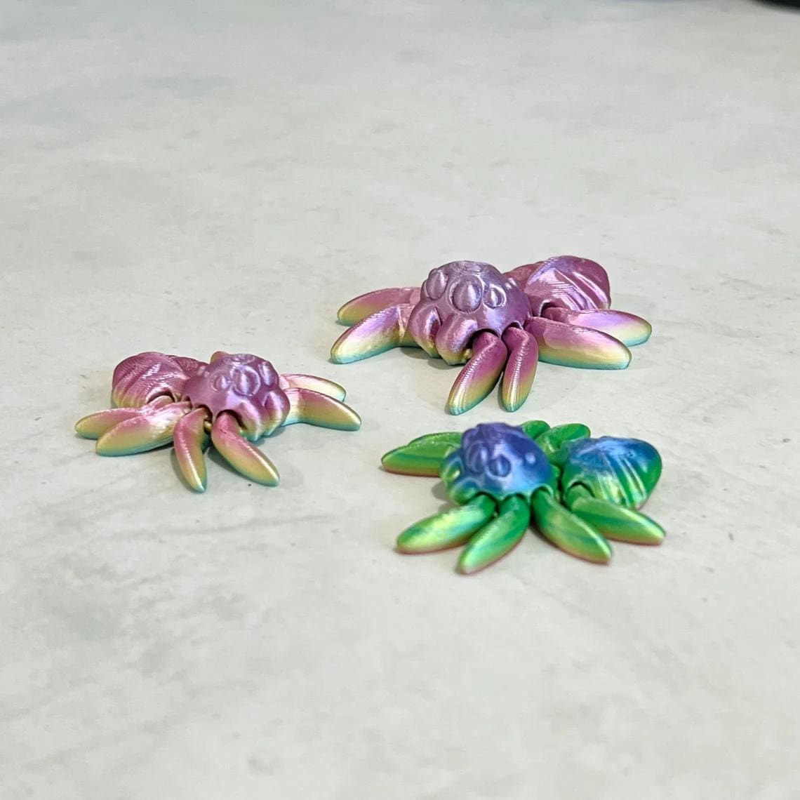 Three colorful, 3D-printed spiders in shades of pink, green, and blue, featuring articulated legs and playful designs.