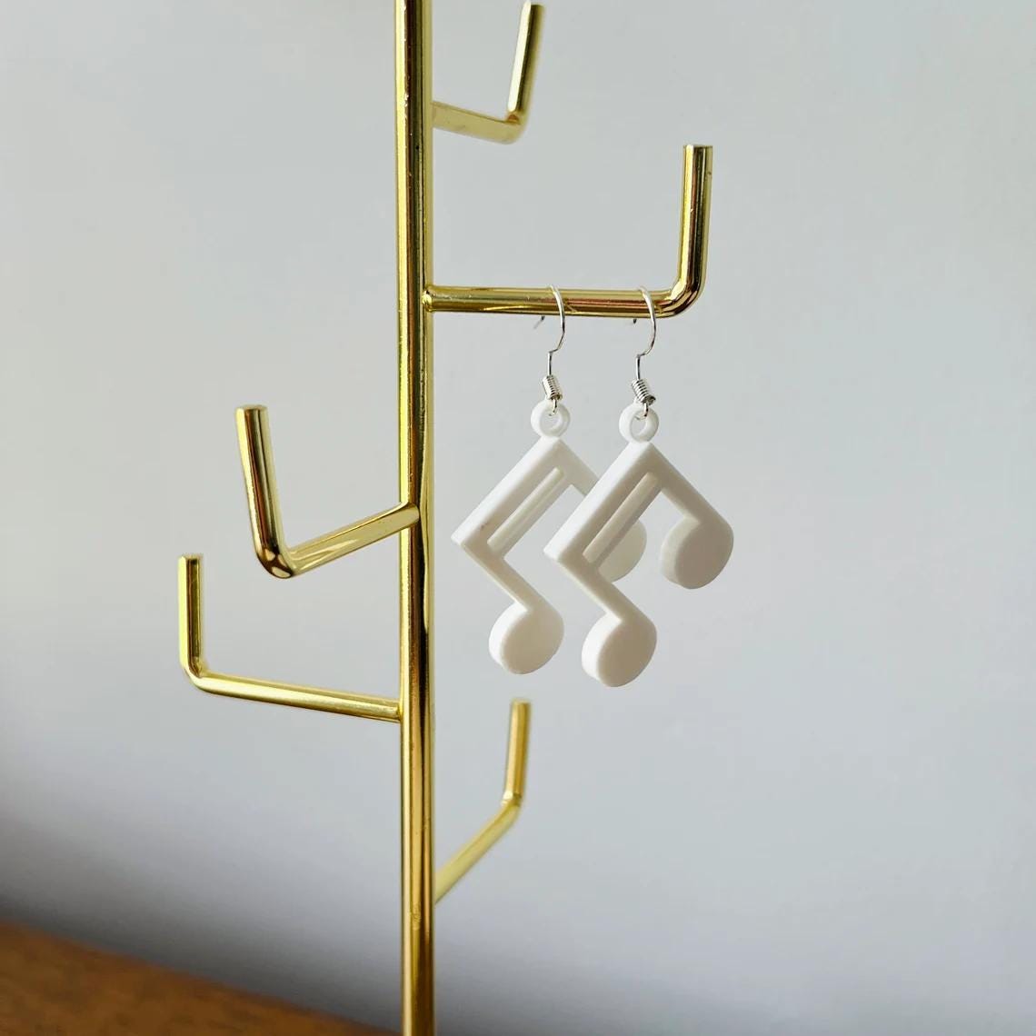 White music note dangle earrings hanging from a gold display stand, showcasing their elegant and lightweight design.