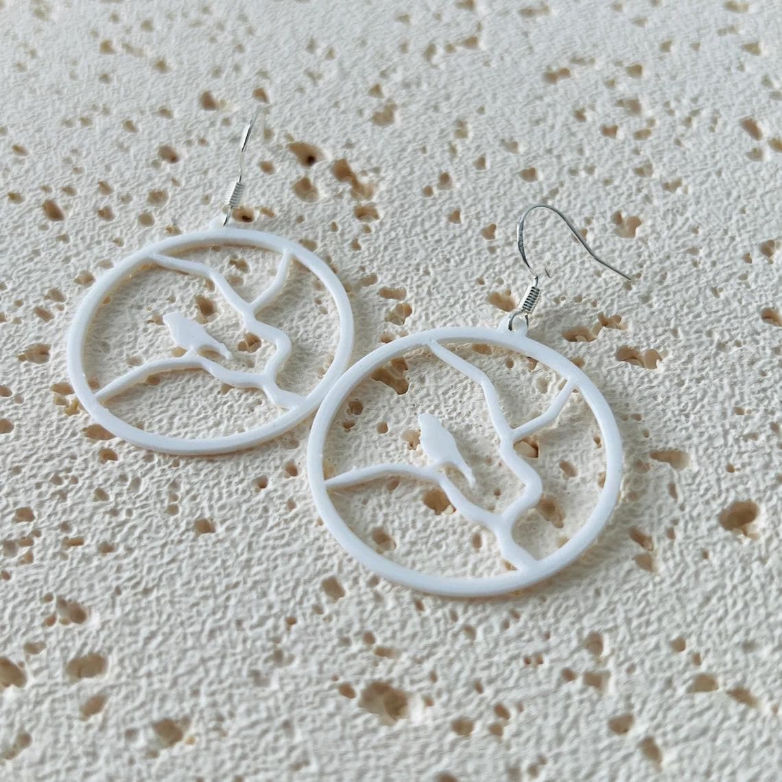 White dangle earrings featuring a bird perched on a branch, crafted from lightweight materials with sterling silver hooks.