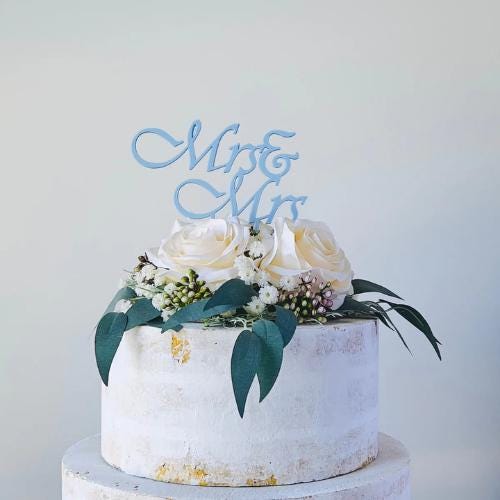 Elegant cake topper featuring "Mrs & Mrs" in soft blue, adorned with white roses and greenery on a textured cake.