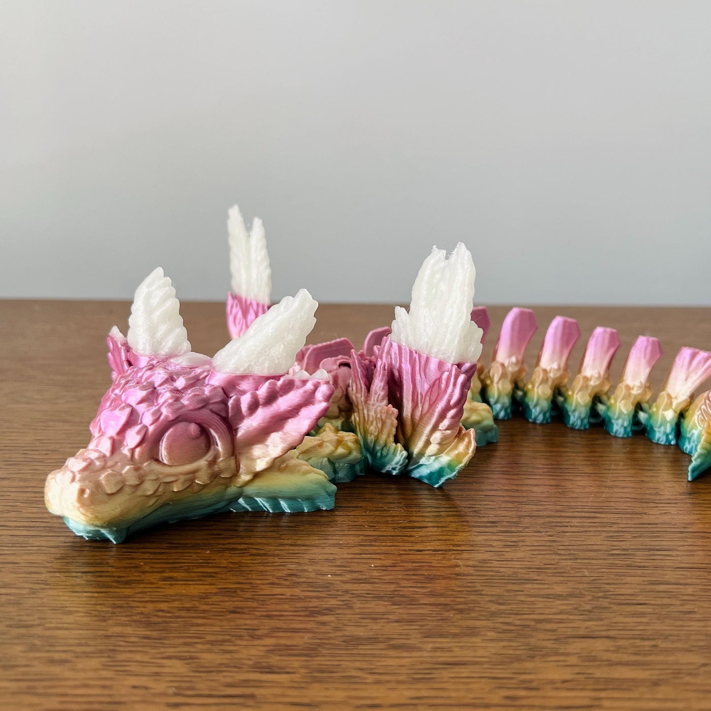 Whimsical dragon with axolotl features, elegant wings, and vibrant pink and blue hues, perched on a wooden surface.