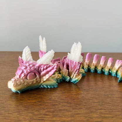 Whimsical dragon with axolotl features, elegant wings, and vibrant pink and blue hues, perched on a wooden surface.