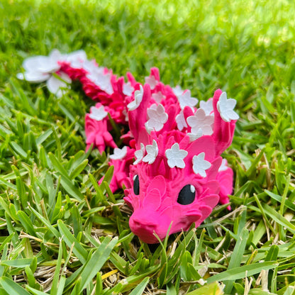 A vibrant pink dragon adorned with delicate white cherry blossom petals, resting on lush green grass.