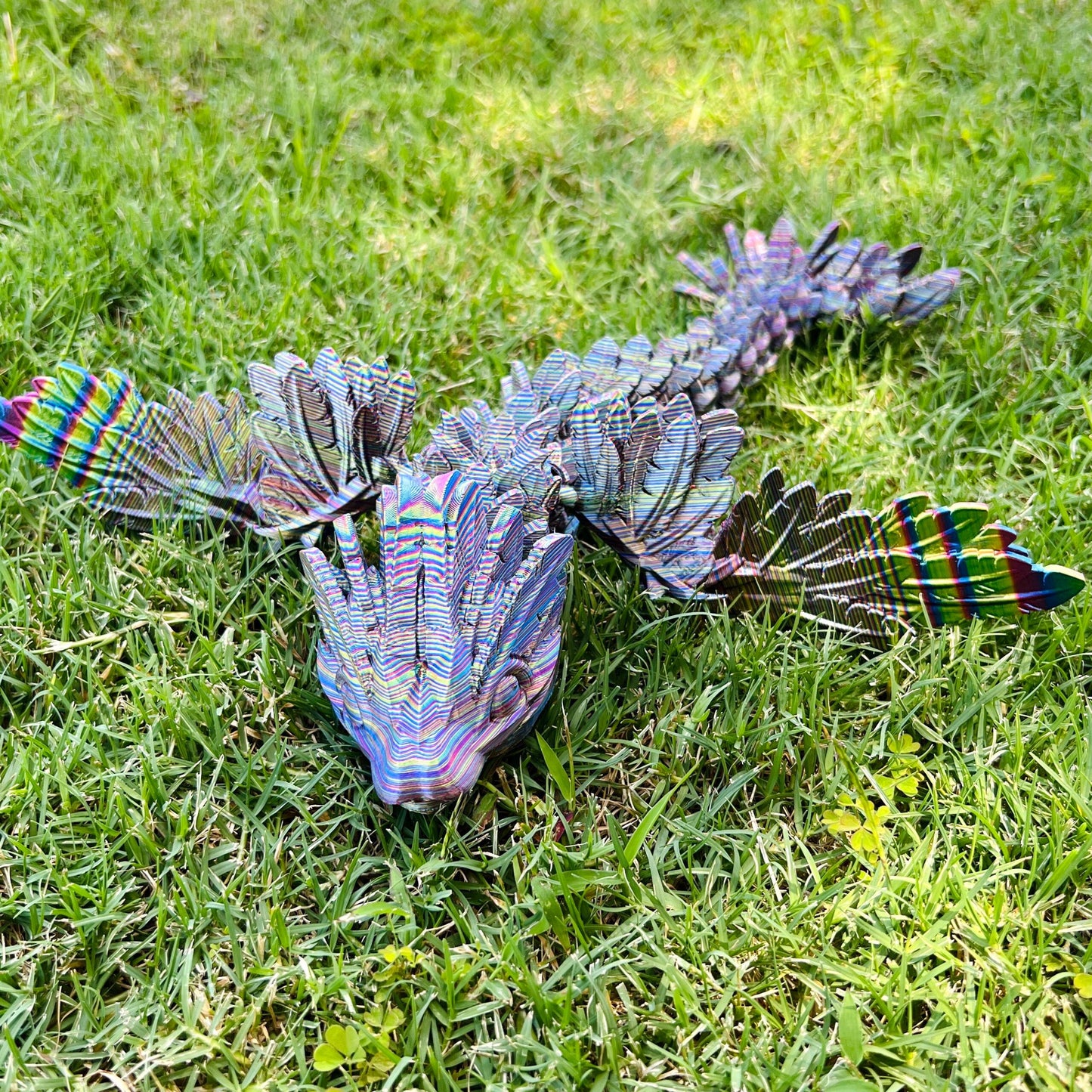 A sleek winged dragon with vibrant, iridescent scales and intricate feathered wings rests on lush green grass.