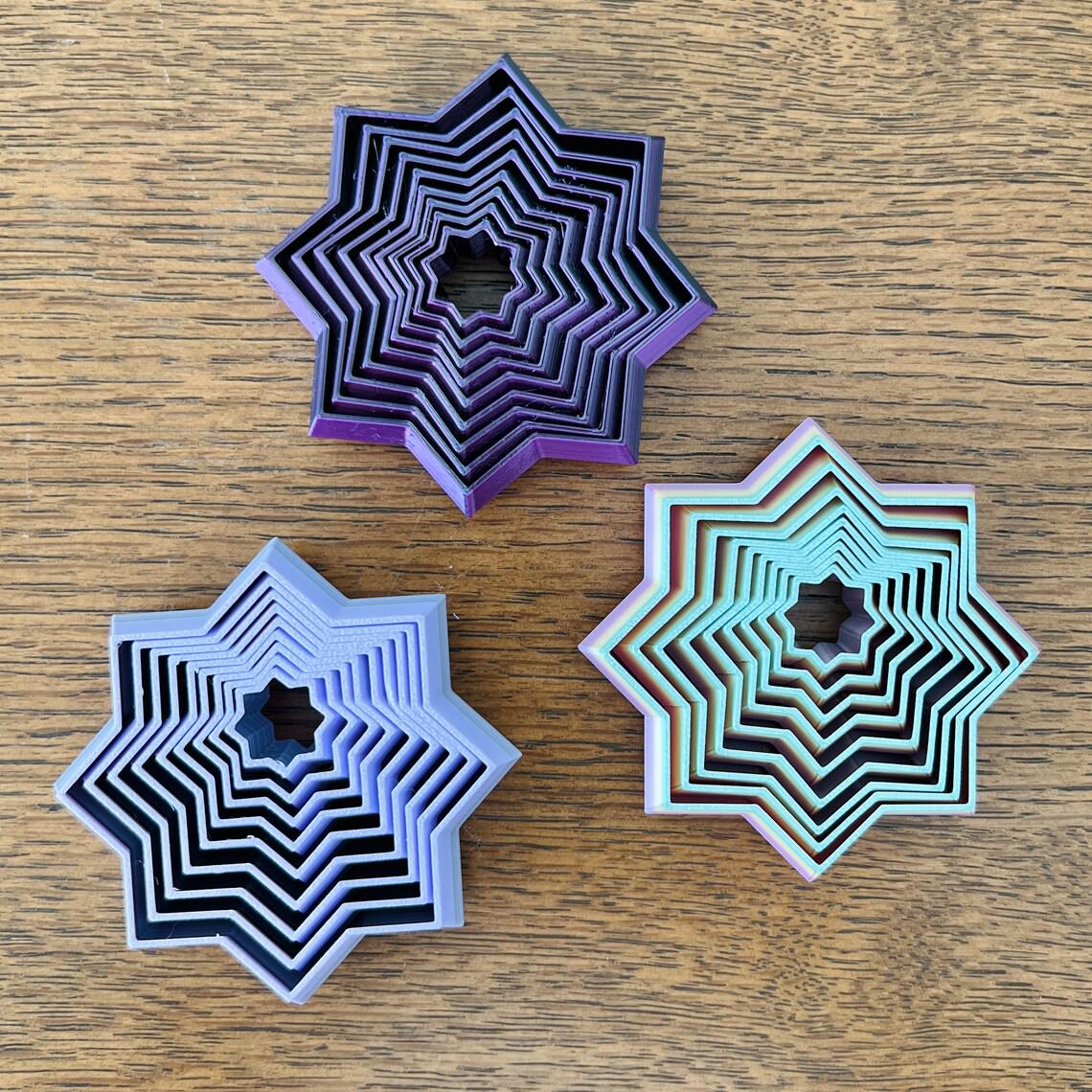 Three colorful star-shaped fidgets with layered designs in purple, teal, and black, resting on a wooden surface.