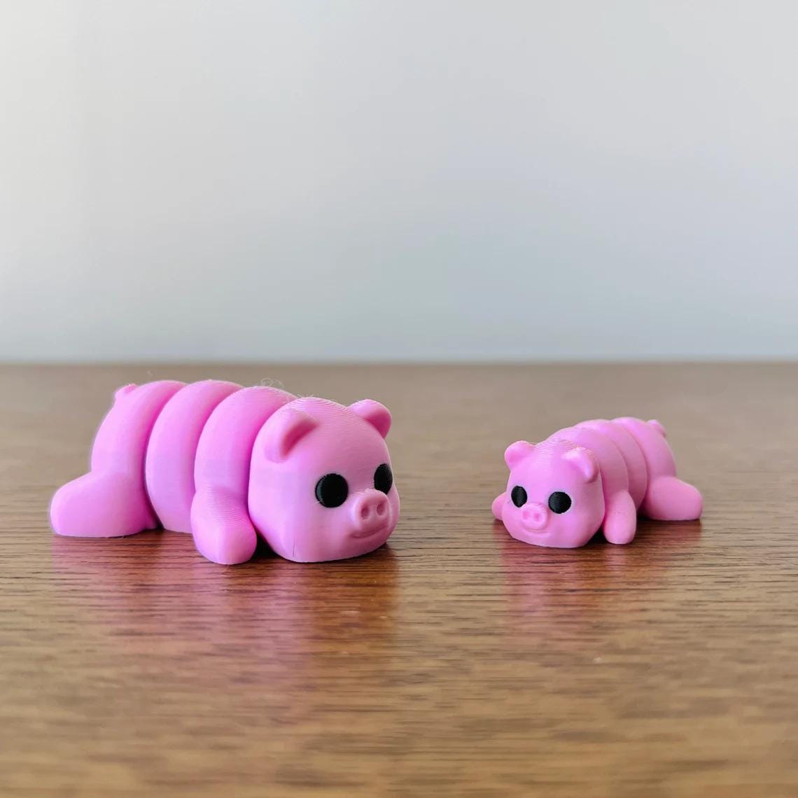 Two adorable pink articulated pigs, one larger and one smaller, resting on a wooden surface with a light background.