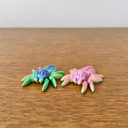 Two colorful, articulated spiders in green-blue and pink hues, resting on a wooden surface. Perfect for sensory play.