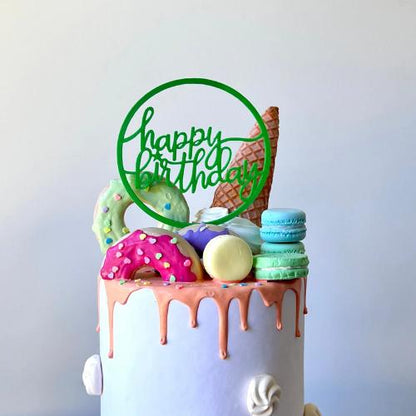 Large green "Happy Birthday" topper on a cake decorated with colorful donuts, macarons, and an ice cream cone.