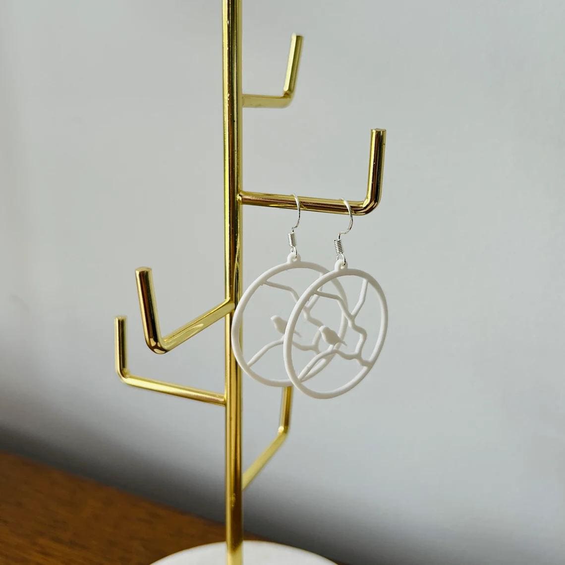White dangle earrings featuring a bird perched on a branch, hanging elegantly from a golden display stand.