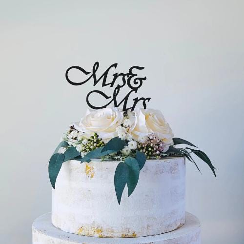 Elegant black "Mrs & Mrs" topper atop a white floral arrangement with roses and greenery on a textured cake surface.