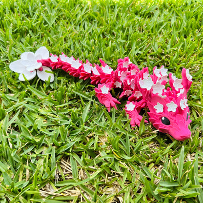 A vibrant pink dragon adorned with delicate white cherry blossoms, resting gracefully on lush green grass.