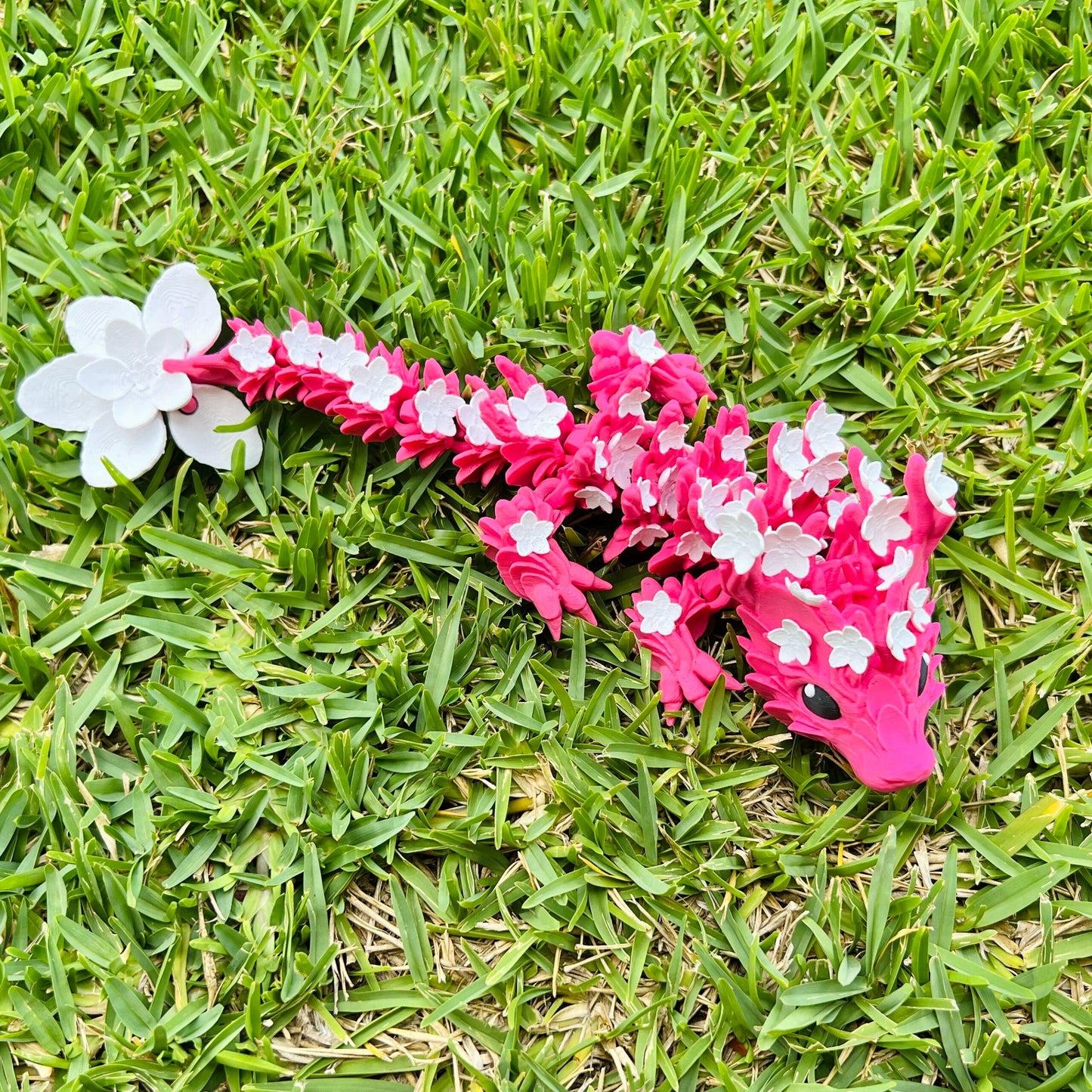 A vibrant pink dragon adorned with delicate white cherry blossoms, resting on a lush green grass background.
